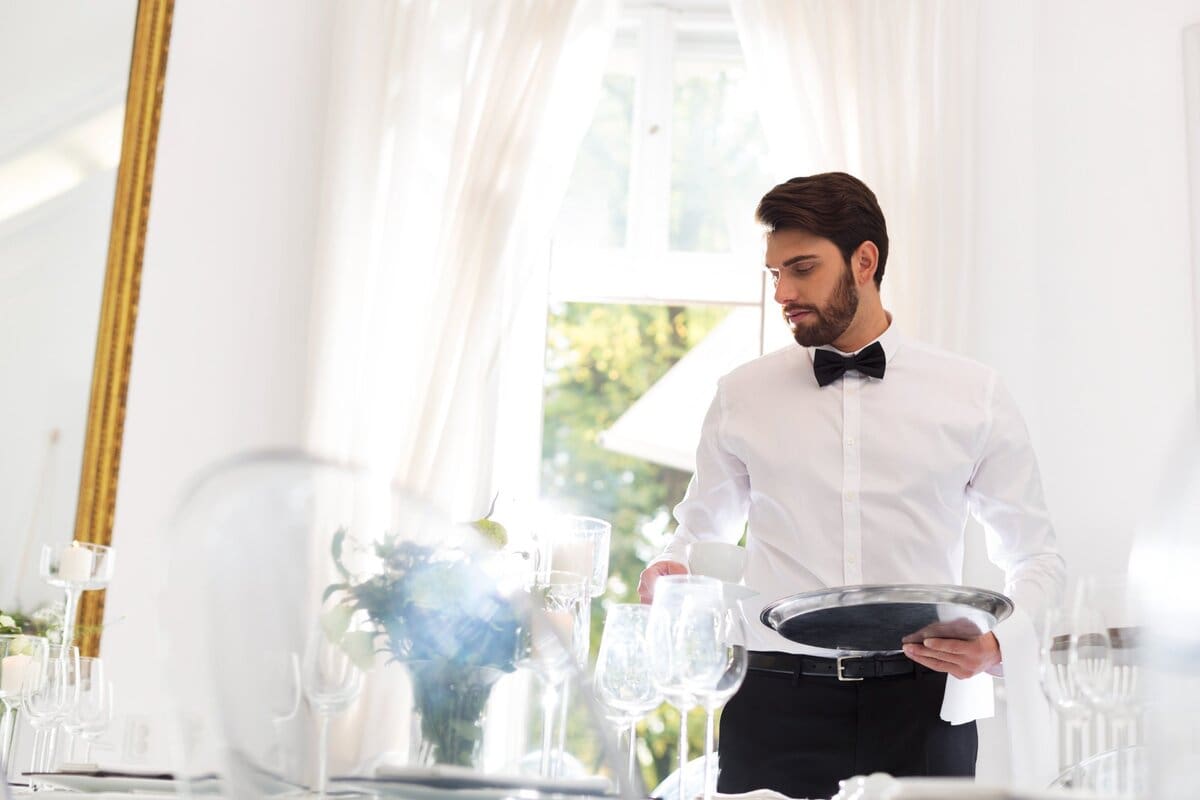A well-dressed male waiter in a white shirt and black bow tie holding a silver tray while arranging glassware on an elegantly set dining table in a bright, upscale restaurant.